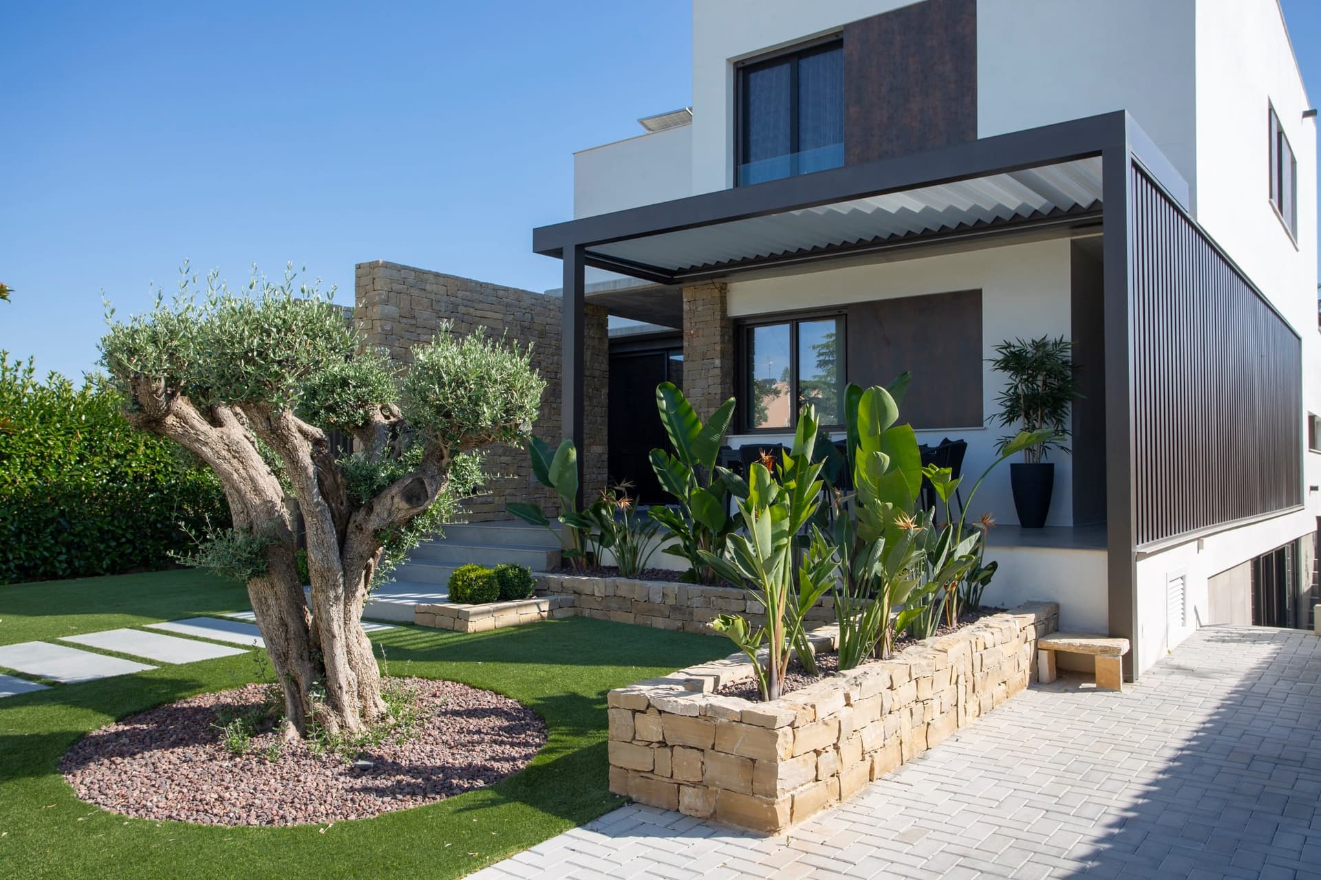 Anthracite bioclimatic pergola over the terrace of a house, with an olive tree in the garden
