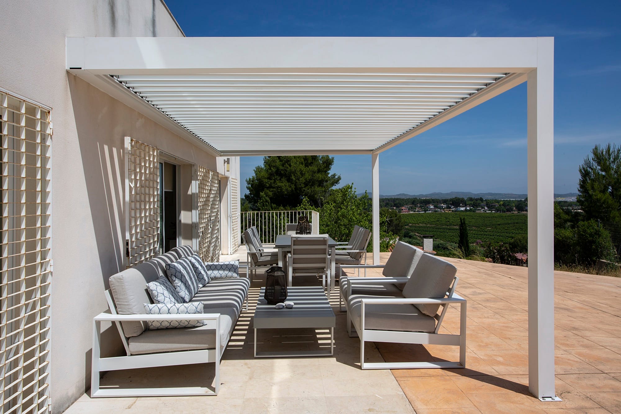Pérgola bioclimática branca sobre um terraço com sofás, com vista para o campo